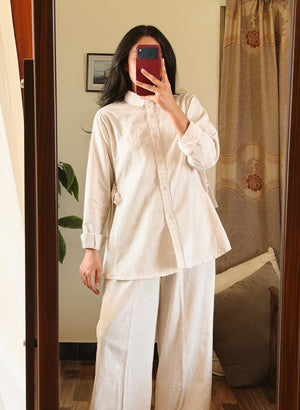 A woman takes a mirror selfie wearing a light beige two-piece linen set consisting of a long-sleeve button-up shirt and matching wide-leg trousers, in a bedroom setting.