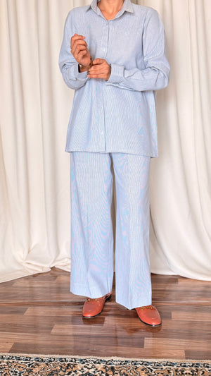 Person wearing a light blue striped shirt and pants standing against a white curtain background.