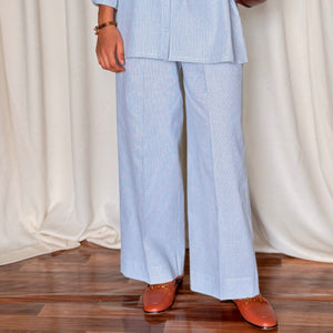 Person wearing a light blue outfit with wide-leg pants and holding a brown bag against a white curtain background.