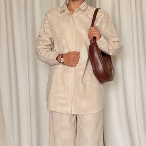 Person wearing a beige striped shirt and pants holding a brown leather bag against a white curtain background.
