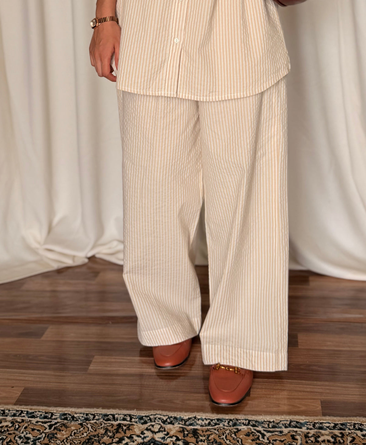 Person wearing a beige striped shirt and pants with a brown handbag against a white curtain background.