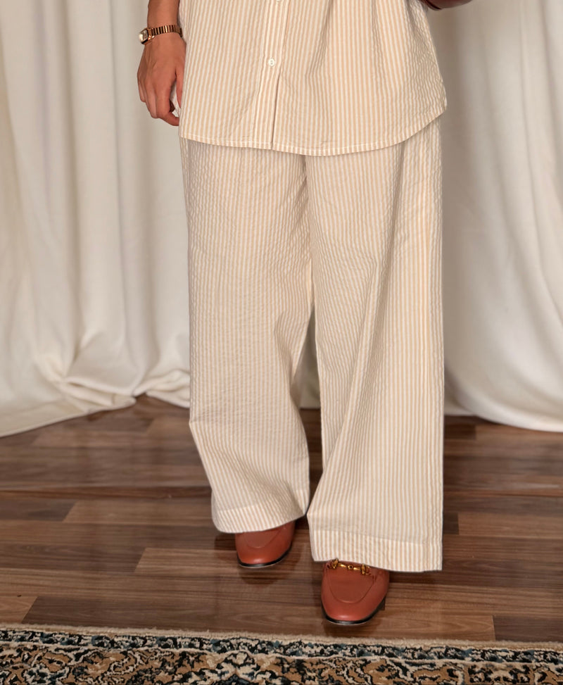 Person wearing a beige striped shirt and pants with a brown handbag against a white curtain background.