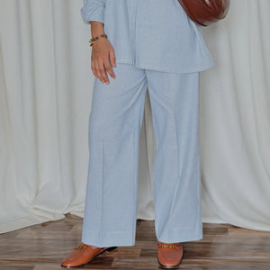 Person wearing a light blue outfit with a brown bag against a white curtain background