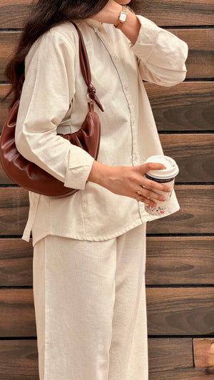 A woman wearing a cream-colored linen two-piece set consisting of a long-sleeve button-up shirt and matching trousers, styled with a brown leather shoulder bag and holding a paper coffee cup against a dark wood plank background.