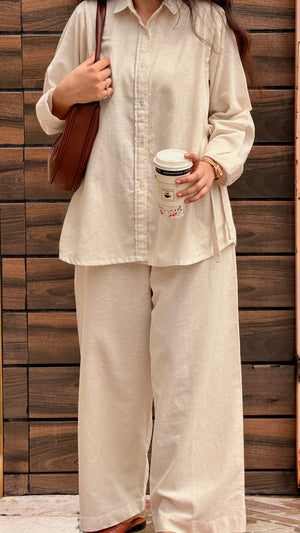 A woman wearing a matching beige linen button-up shirt and wide-leg pants set, holding a brown paper coffee cup and carrying a brown leather shoulder bag against a dark wood plank wall.