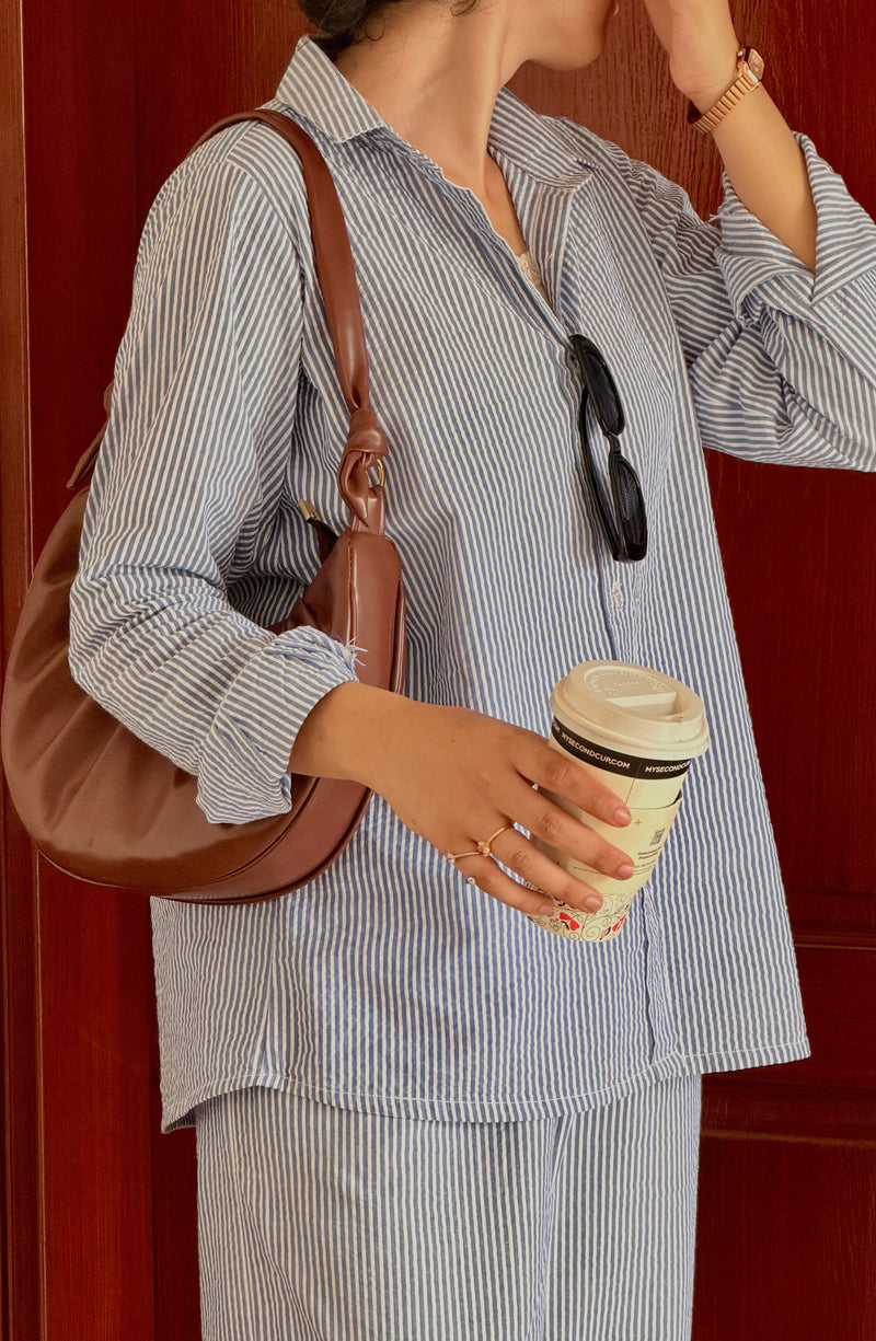 A person wearing a blue and white vertically striped button-down shirt and matching striped pants, holding a takeaway coffee cup and wearing a brown shoulder bag, standing against a dark reddish-brown wooden door.