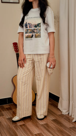 A woman wearing a white graphic t-shirt and beige pinstriped wide-leg trousers, standing in a room with a wooden floor, a guitar, and a curtain.