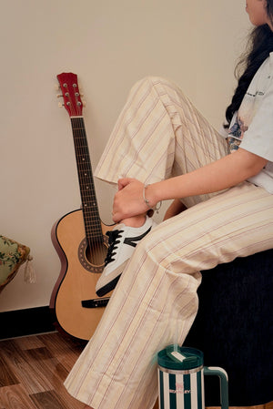 A person sitting on a dark stool wearing beige and white vertically striped wide-leg pants and white sneakers, with an acoustic guitar and a striped tumbler nearby in a styled indoor setting.
