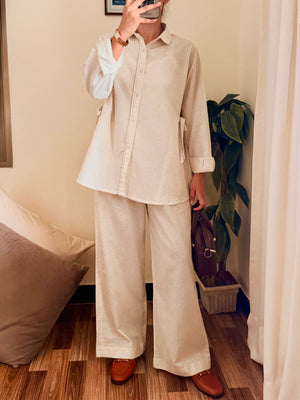 A person takes a mirror selfie wearing a matching beige linen two-piece set consisting of a long-sleeve button-up shirt and wide-leg trousers, paired with brown leather loafers in a home setting.