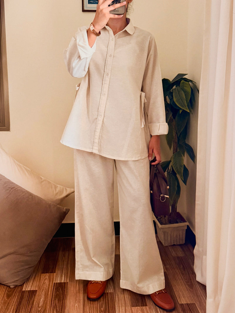 A person takes a mirror selfie wearing a matching beige linen two-piece set consisting of a long-sleeve button-up shirt and wide-leg trousers, paired with brown leather loafers in a home setting.