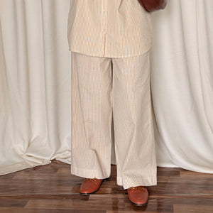 Person wearing a beige striped shirt and pants holding a brown leather bag against a white curtain background.