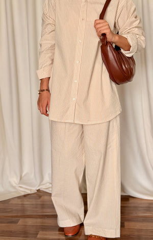 Person wearing a beige striped outfit with a brown handbag against a white curtain background