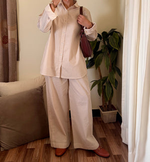 Person taking a mirror selfie wearing a beige outfit in a room with a plant and curtain.