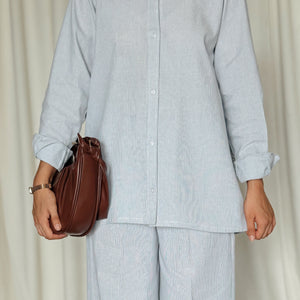 Person wearing a light blue outfit with wide-leg pants and holding a brown bag against a neutral background.
