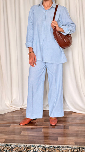 Person wearing a light blue outfit with wide-leg pants and holding a brown bag against a white curtain background.