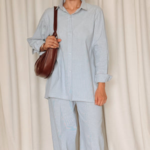 Person wearing a light blue outfit with brown shoes and a bag, standing against a white curtain.