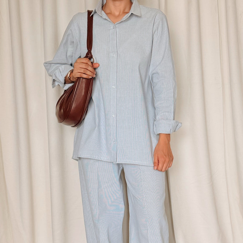 Person wearing a light blue outfit with brown shoes and a bag, standing against a white curtain.