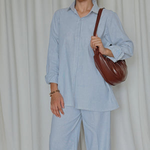 Person wearing a light blue outfit with a brown bag against a white curtain background