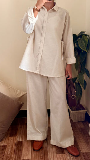 Person wearing a beige shirt and white pants taking a mirror selfie in a room with wooden flooring and a plant.