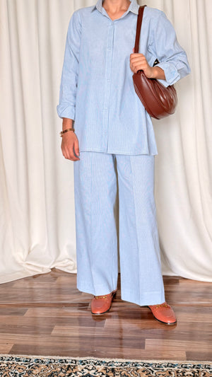 Person wearing a light blue outfit with wide-leg pants and holding a brown bag on a wooden floor.