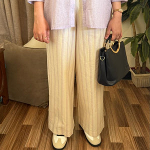 Person wearing a light-colored outfit with wide-leg pants and holding a black handbag indoors.