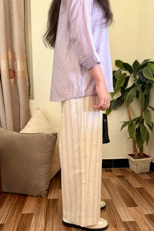 Person wearing a light-colored shirt and beige pants standing in a room with wooden flooring and a plant.
