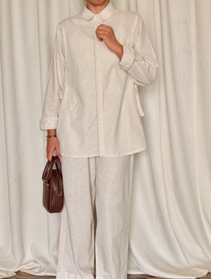 Person wearing a light-colored outfit with a brown bag against a white curtain background