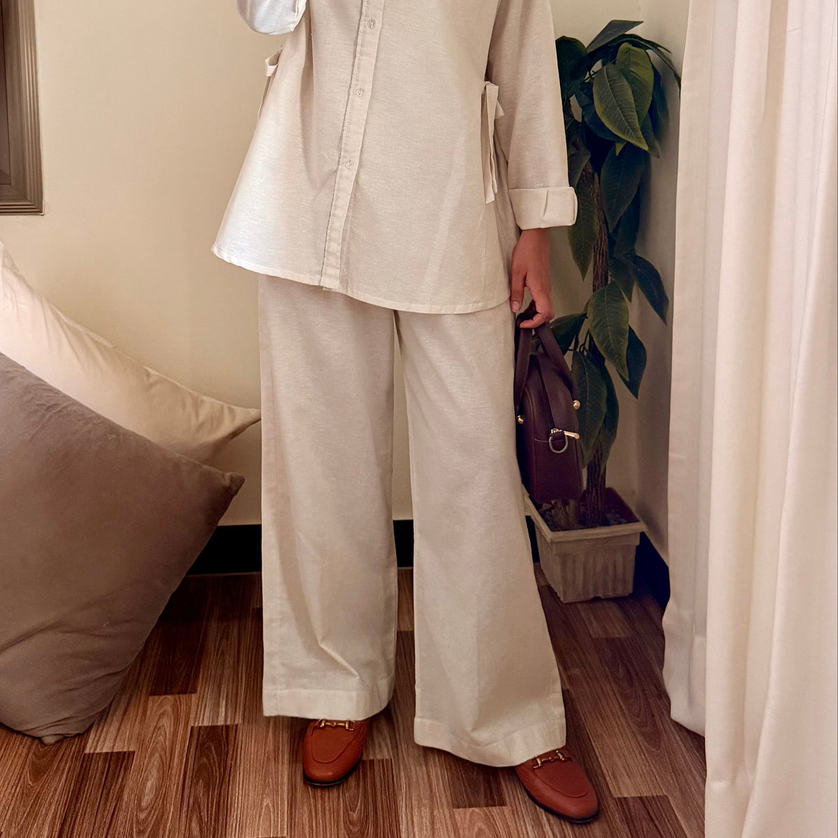 Person wearing a beige shirt and white pants taking a mirror selfie in a room with wooden flooring and a plant.
