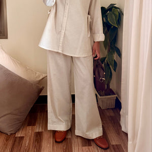 Person wearing a beige shirt and white pants taking a mirror selfie in a room with wooden flooring and a plant.