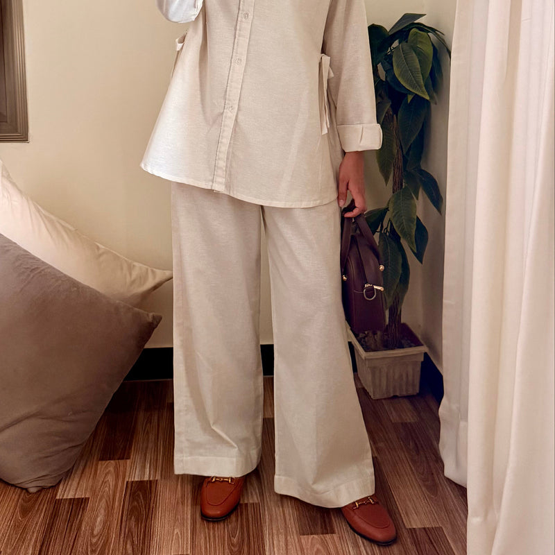 Person wearing a beige shirt and white pants taking a mirror selfie in a room with wooden flooring and a plant.