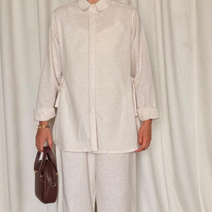 Person wearing a light-colored outfit with wide-leg pants and holding a brown bag against a white curtain background.