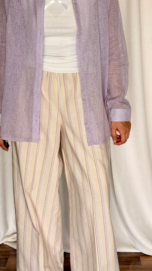 Person wearing a light purple shirt over a white top with beige striped pants against a white curtain background