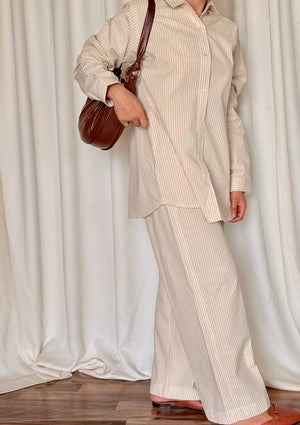 Person wearing a beige striped outfit with a brown bag against a white curtain background