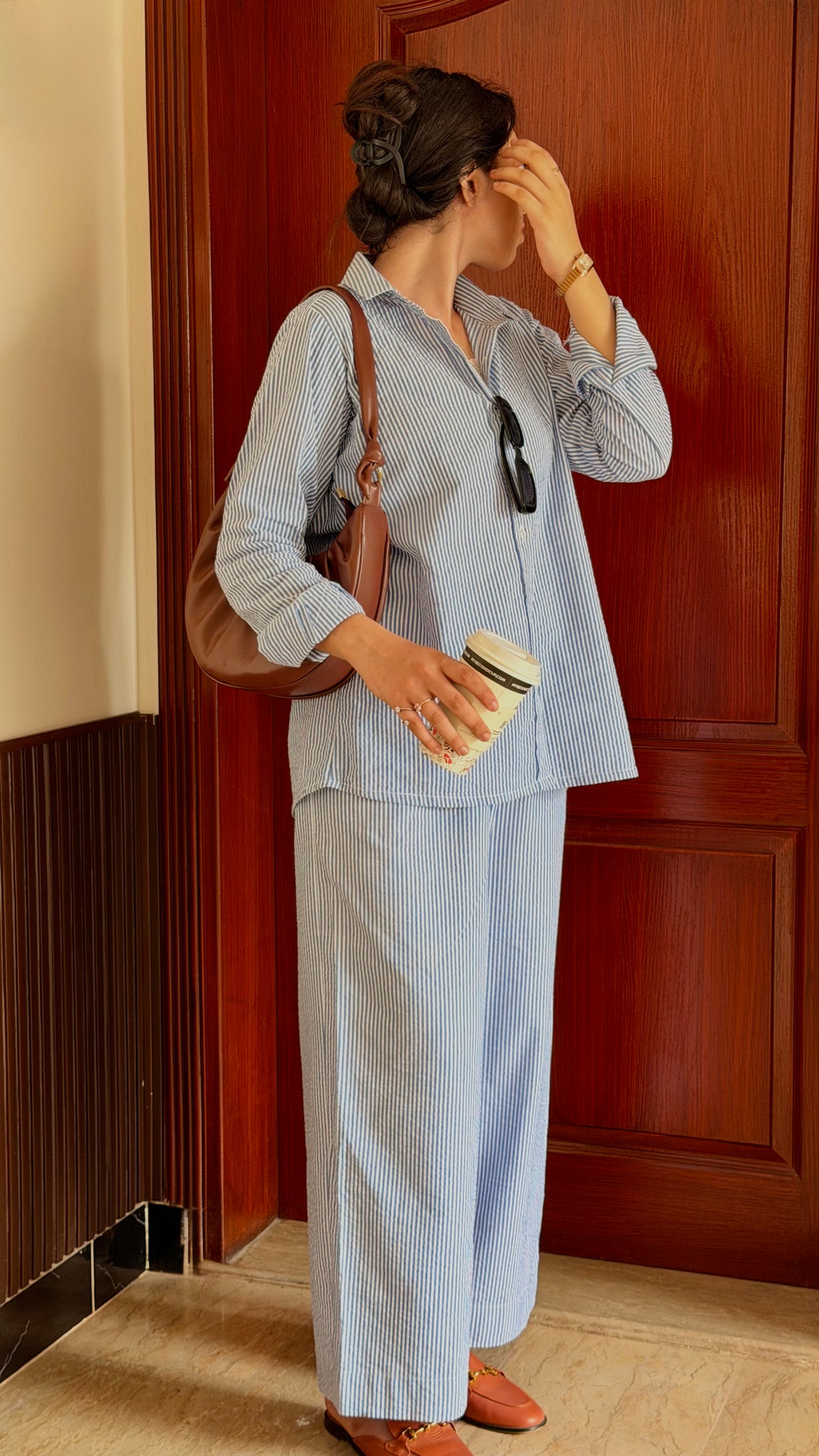 A woman stands in front of a dark wood door wearing a light blue and white striped button-up shirt and matching wide-leg pants. She is holding a white coffee cup and wearing a brown shoulder bag.