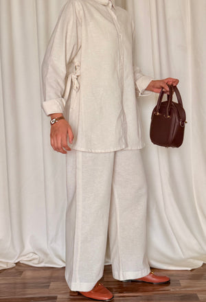 Person wearing a light-colored outfit holding a brown handbag against a white curtain background
