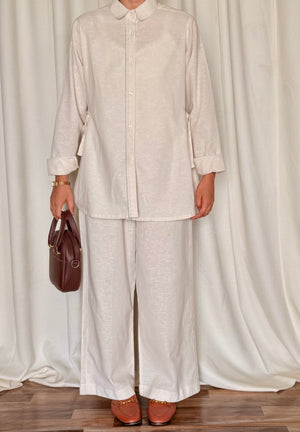Person wearing a light-colored outfit with wide-leg pants and holding a brown bag against a white curtain background.